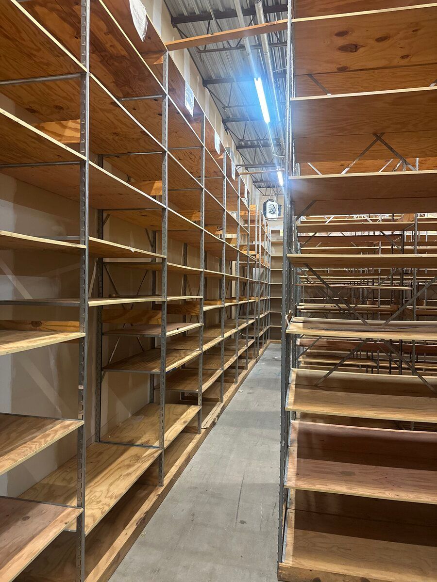 Two long rows of empty wooden industrial shelving units in a storage area with a concrete floor and overhead lighting.