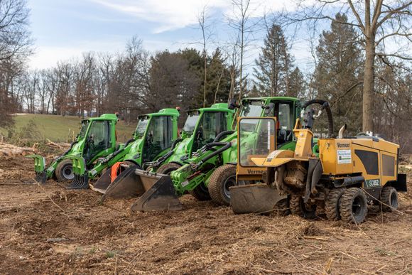 Three green Avant loaders and one yellow stump grinder parked in a row on a dirt field with trees in the background.