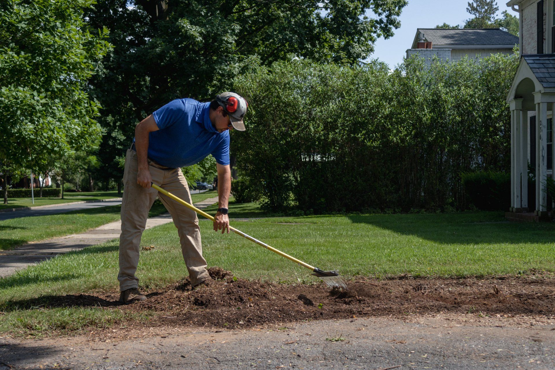 A person in a blue shirt and khaki pants rakes mulch into a garden bed next to a house on a sunny day.