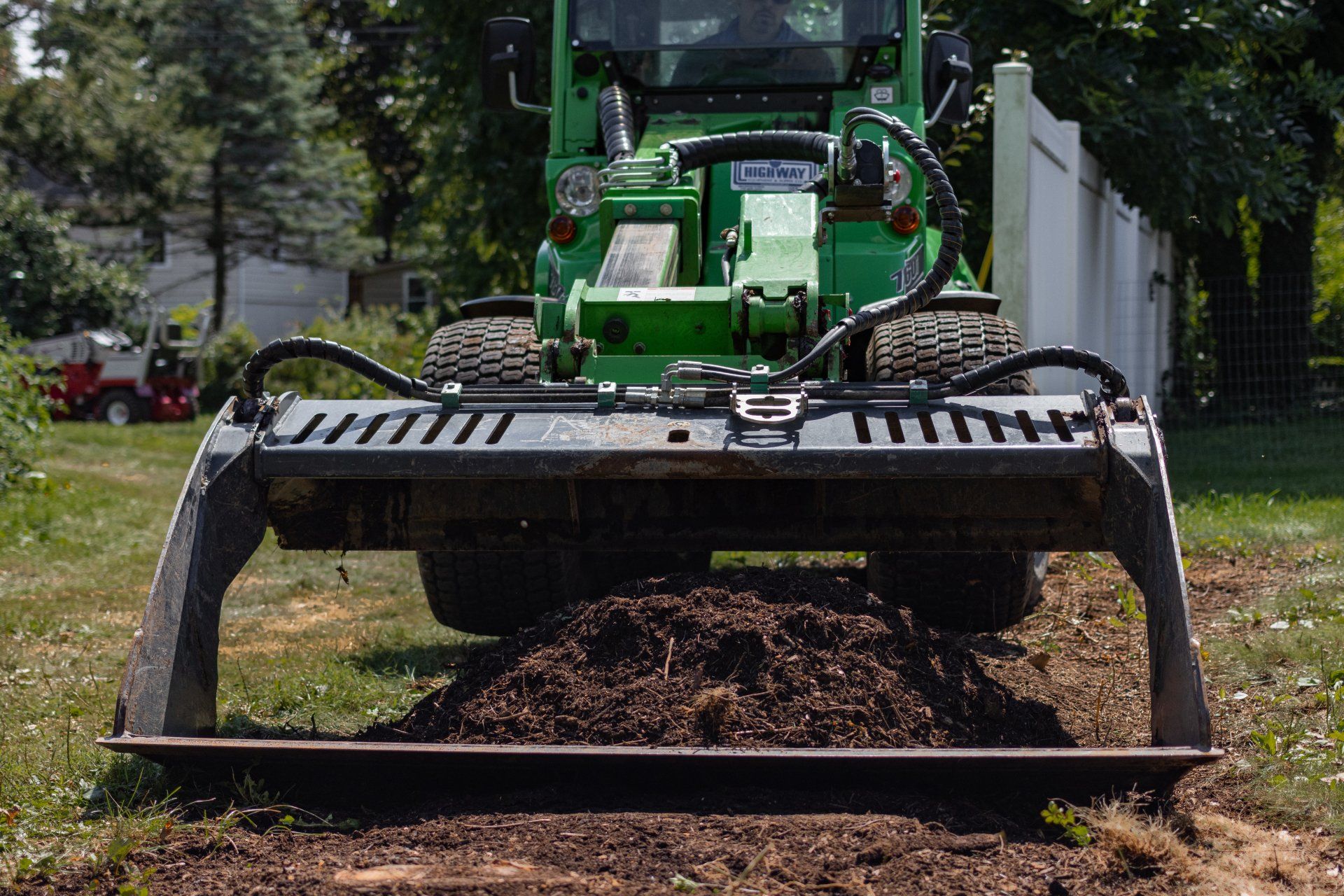 A green utility loader with a grapple bucket moving a pile of dark soil outdoors on a sunny day.