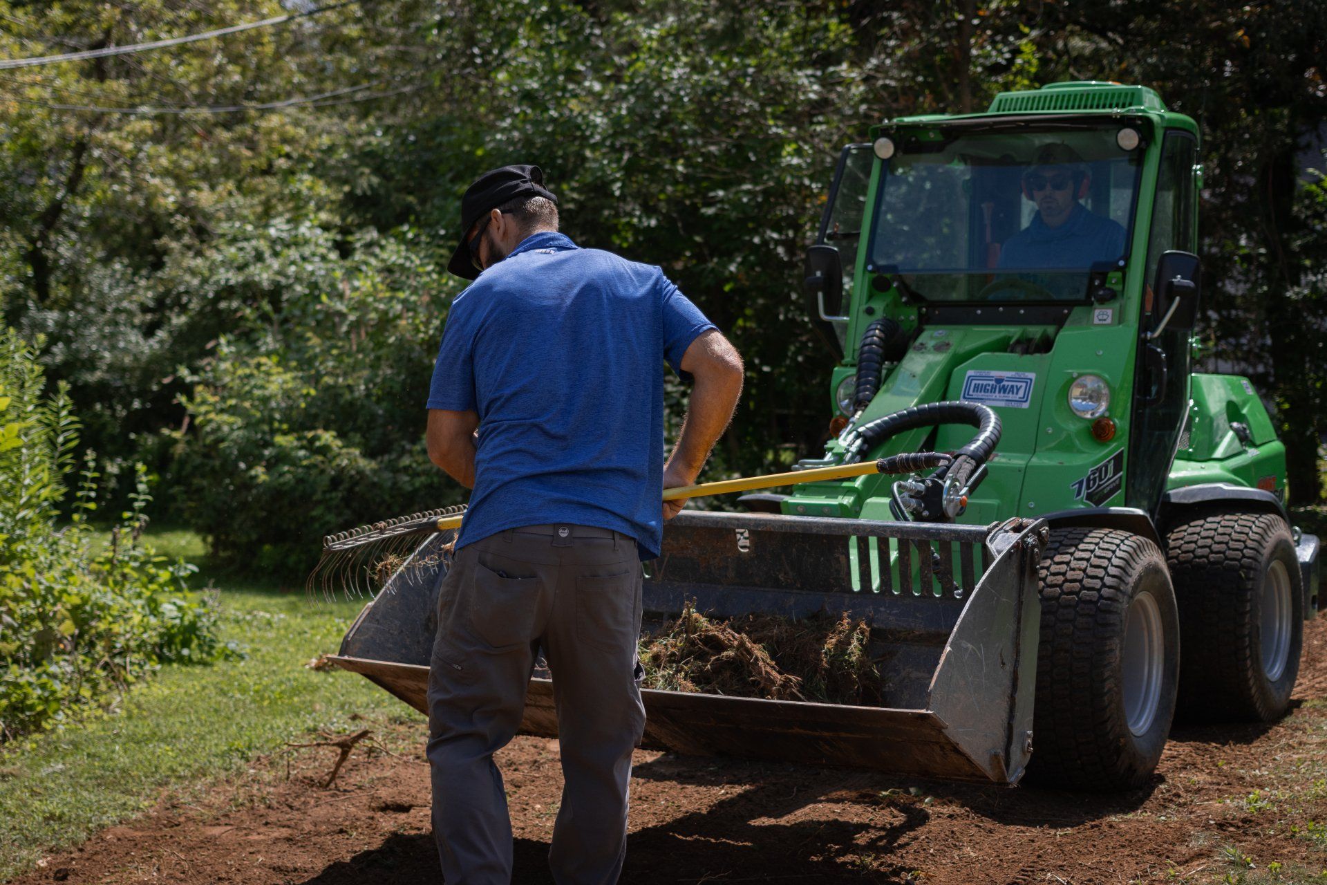 A person in a blue shirt uses a shovel to move soil into the bucket of a green compact loader on a sunny day.