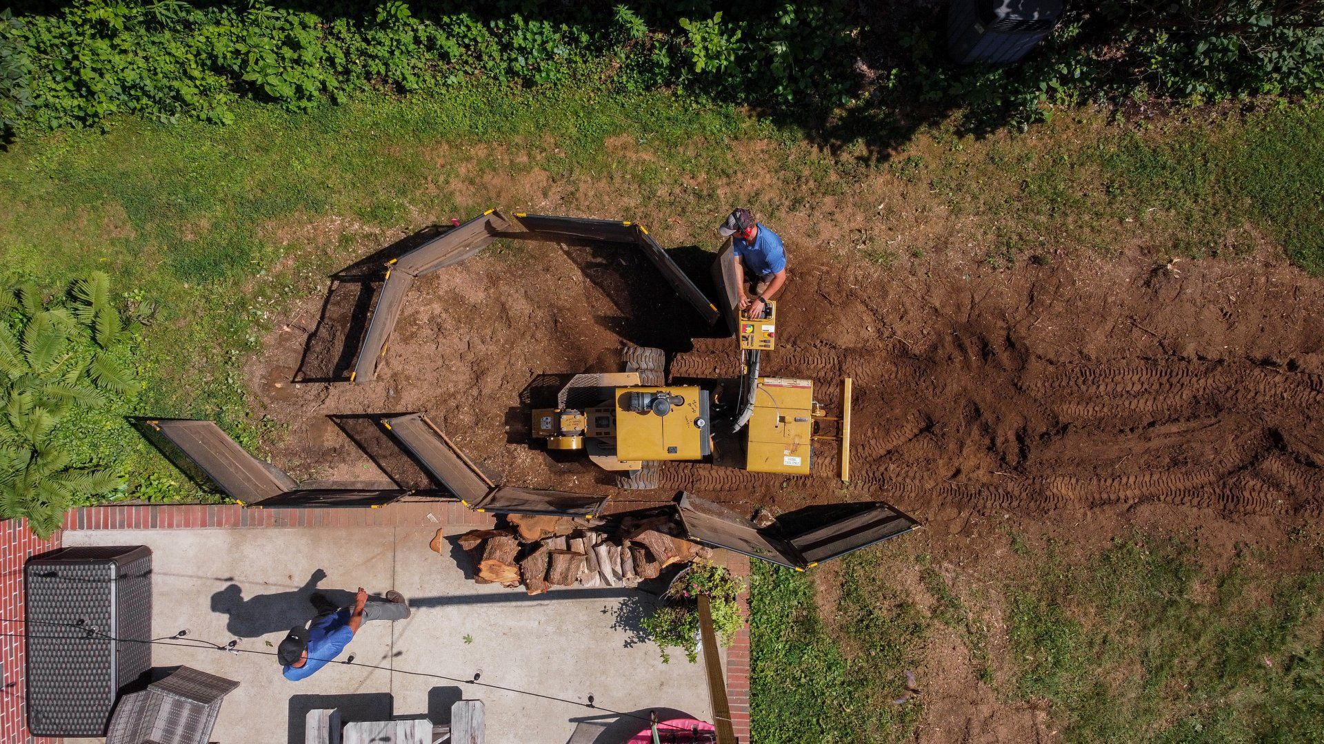 A person operates a yellow trencher to dig in a grassy backyard near a patio and wooden fence.