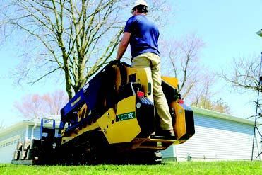 A person stands on the back platform of a yellow and blue Vermeer mini skid steer on a grassy lawn near a house.