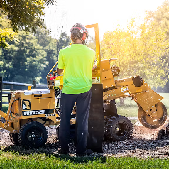A person in a bright neon shirt uses a yellow stump grinder to clear a tree stump outdoors.