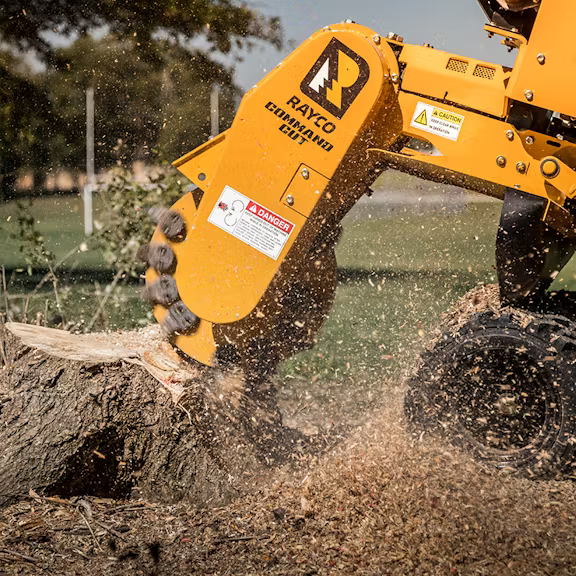 A yellow Rayco stump grinder chewing through a tree stump, creating a spray of wood chips in an outdoor setting.