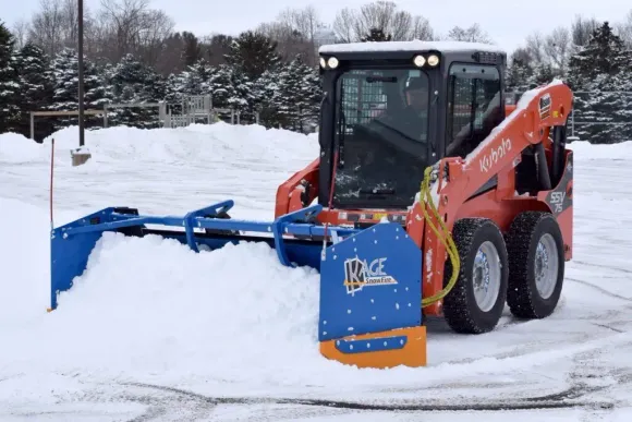 An orange skid steer loader with a large blue snow pusher attachment clearing snow in a snowy outdoor lot.