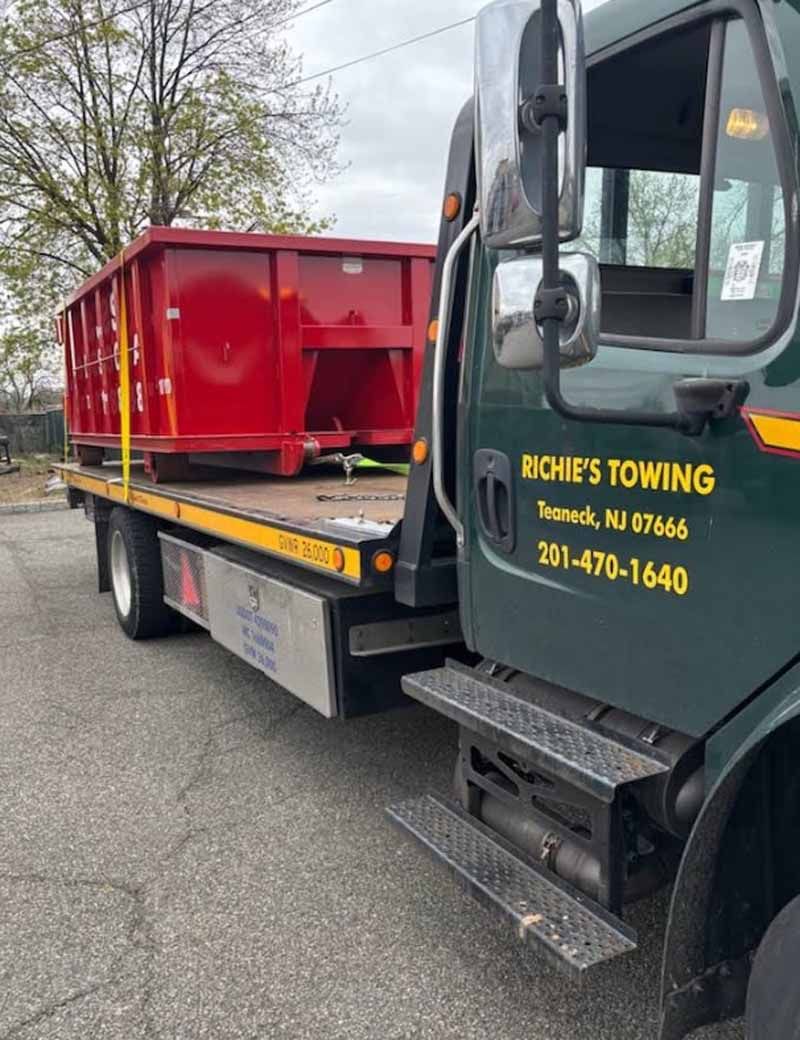 A green towing truck is carrying a red dumpster.