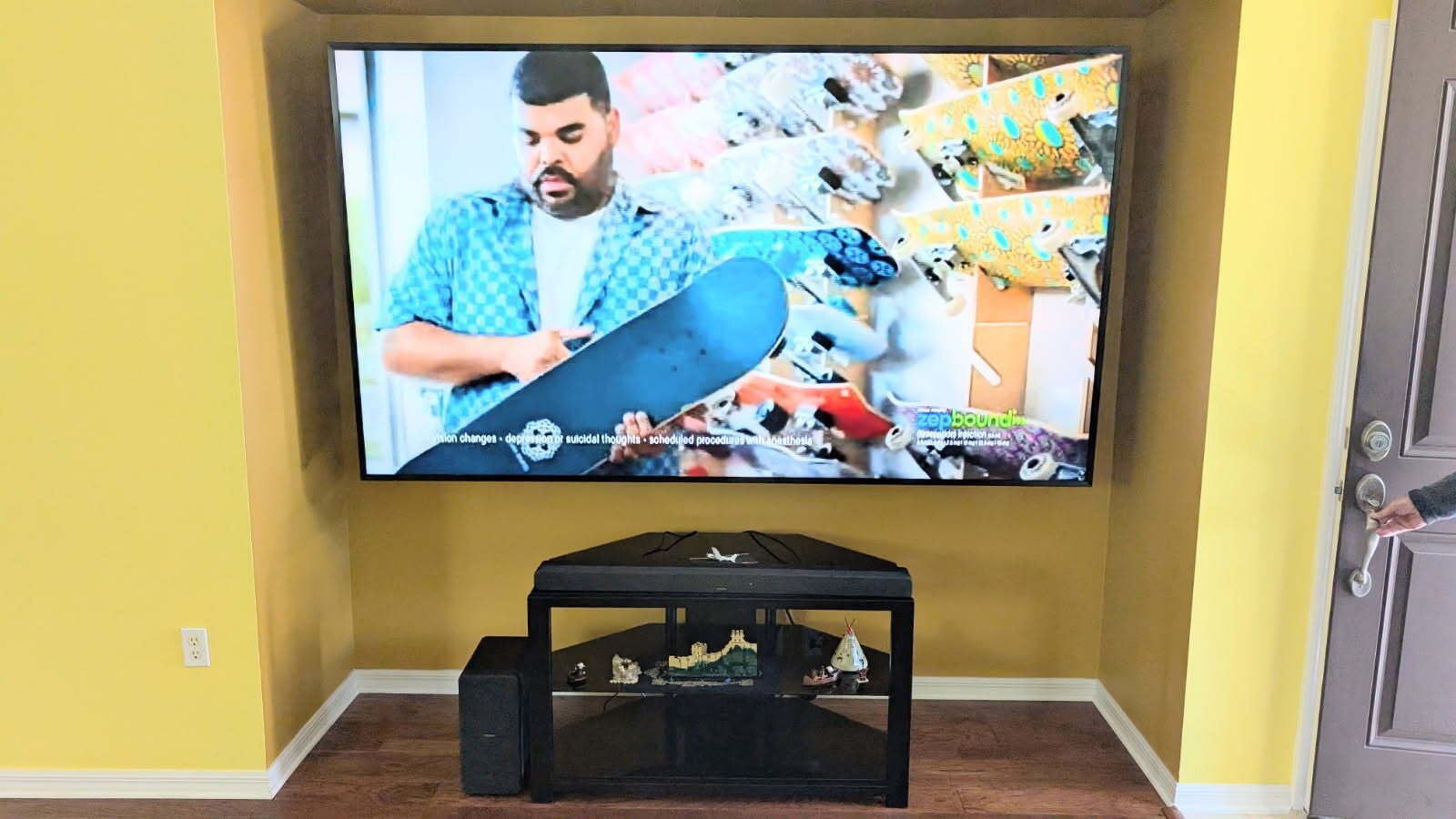 TV in a yellow-walled nook with a man holding a skateboard. Black media console below, and a door on the right.