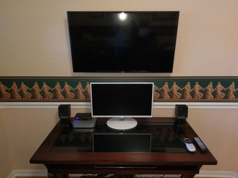 A computer monitor sits on a desk under a flat screen tv