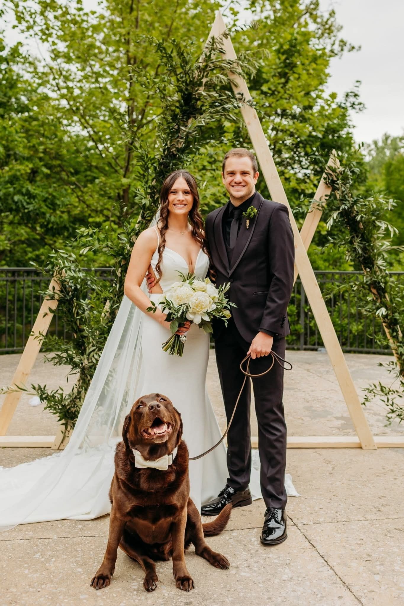 A couple kneels with their chocolate lab in front of a fireplace. They smile, touching the dog.