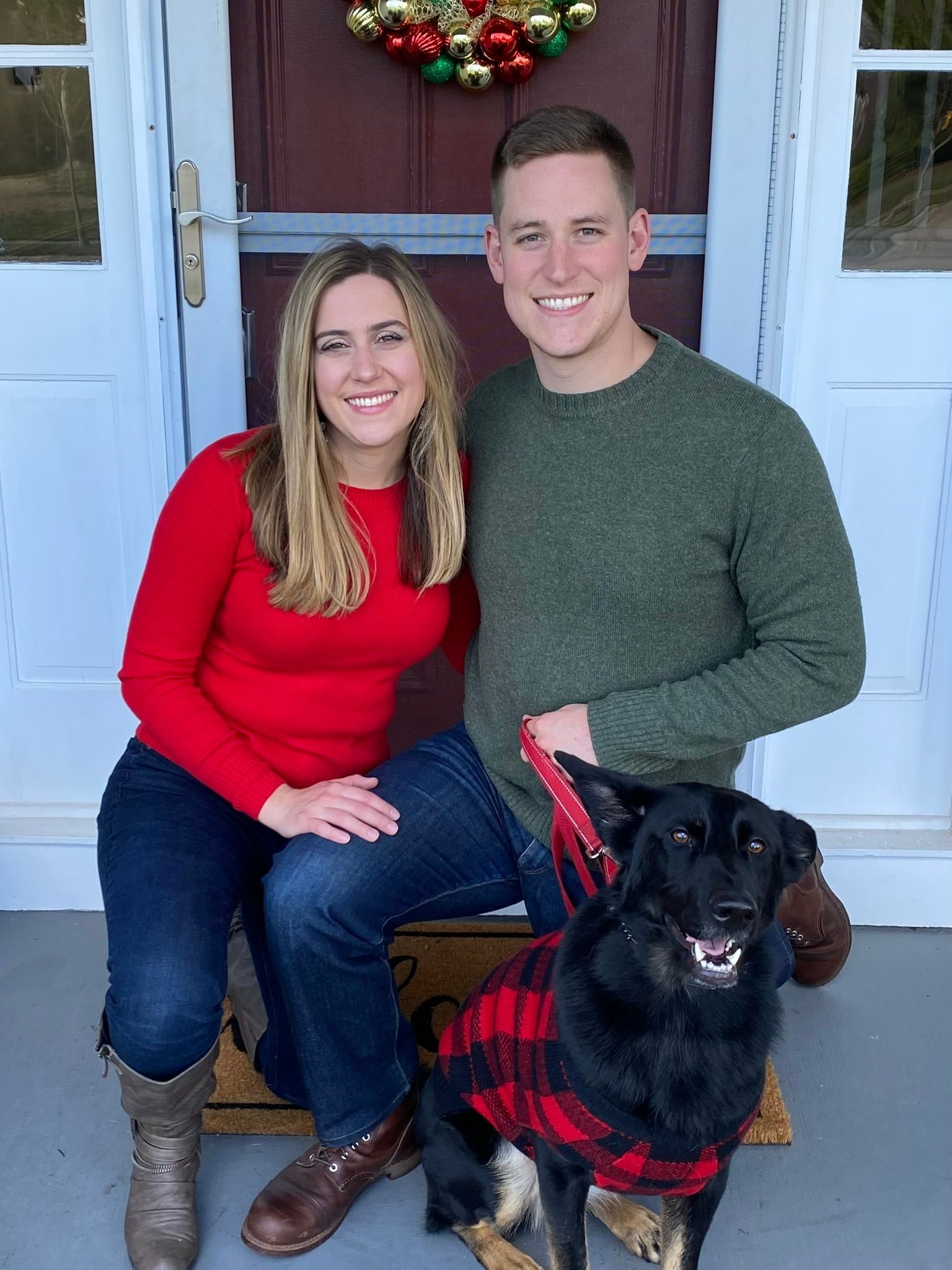 Couple, smiling, poses with dog on front porch. Woman wears red, man in green, dog in red plaid.