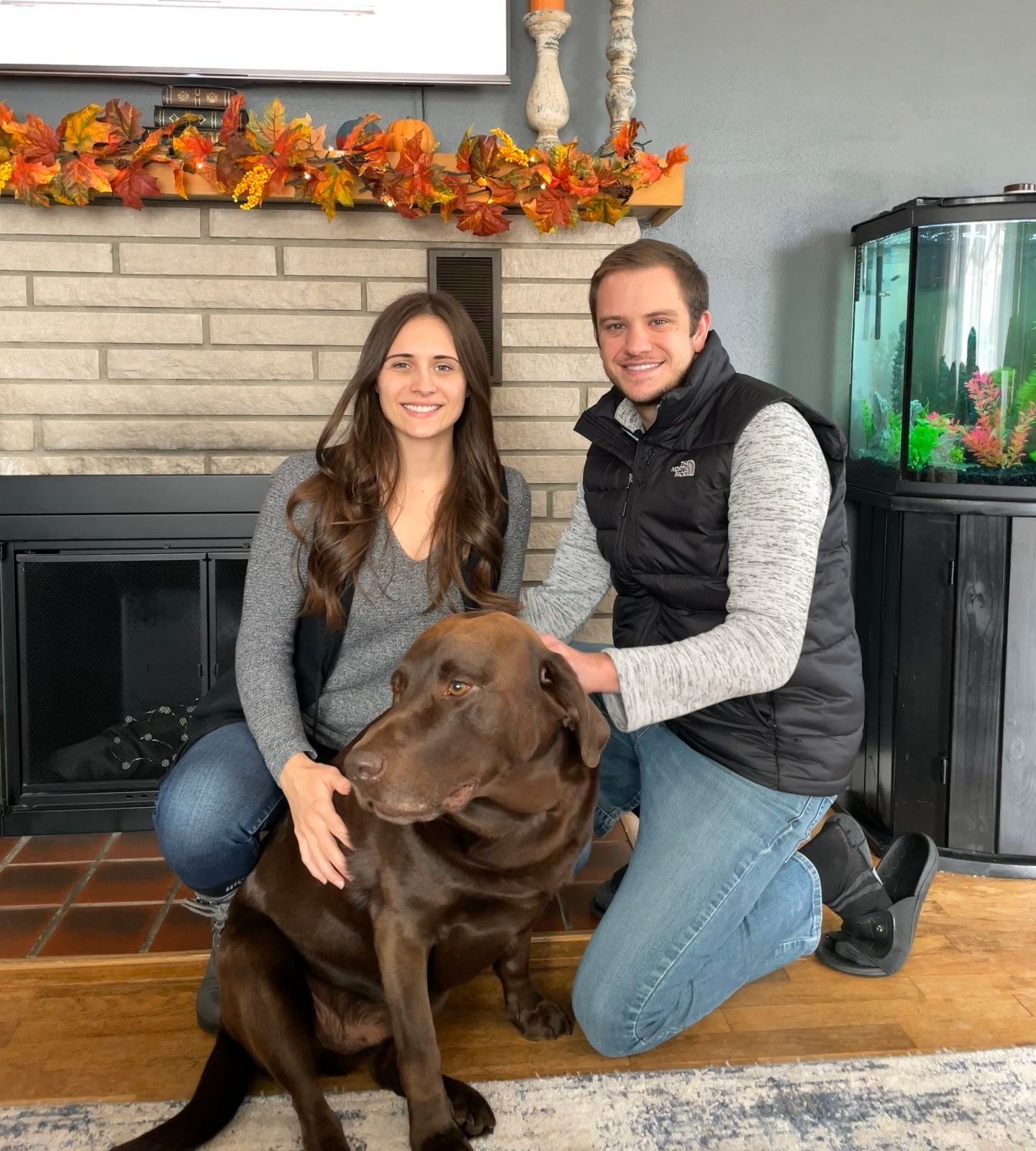 A couple kneels with their chocolate lab in front of a fireplace. They smile, touching the dog.
