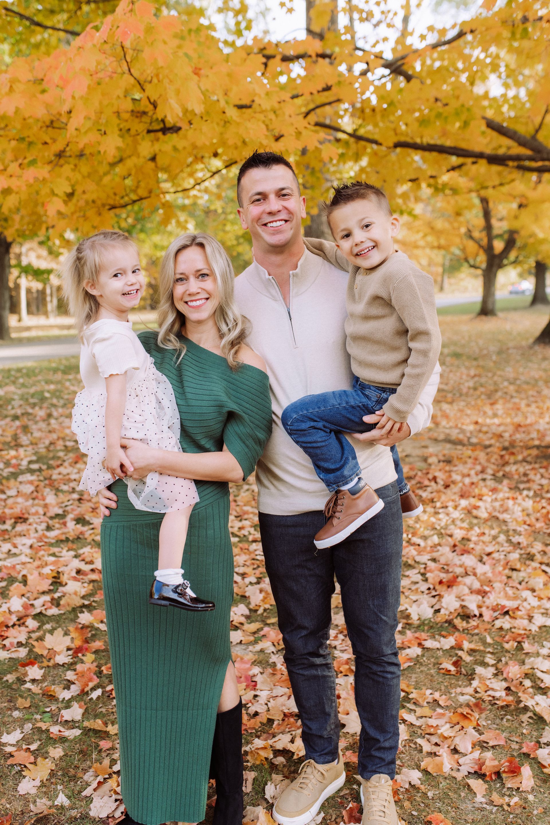 Family of four poses outdoors in front of autumn foliage; mother holds daughter, father holds son.