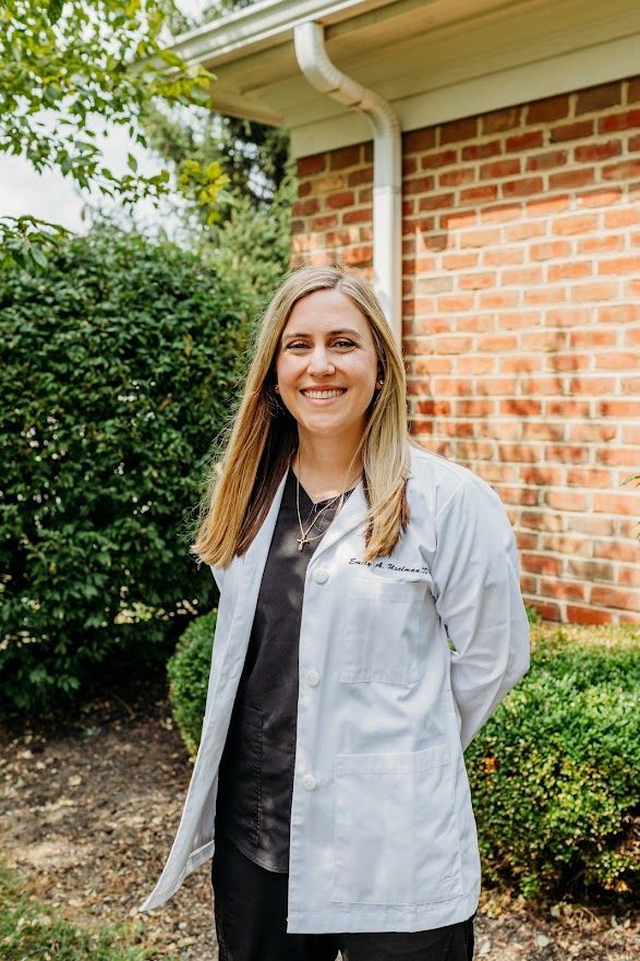 A person in a white medical coat and dark scrubs smiles outdoors against a brick building and greenery.