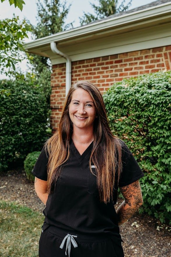 A smiling person with long hair and visible arm tattoos wearing a black scrub set, standing outside by a brick building.