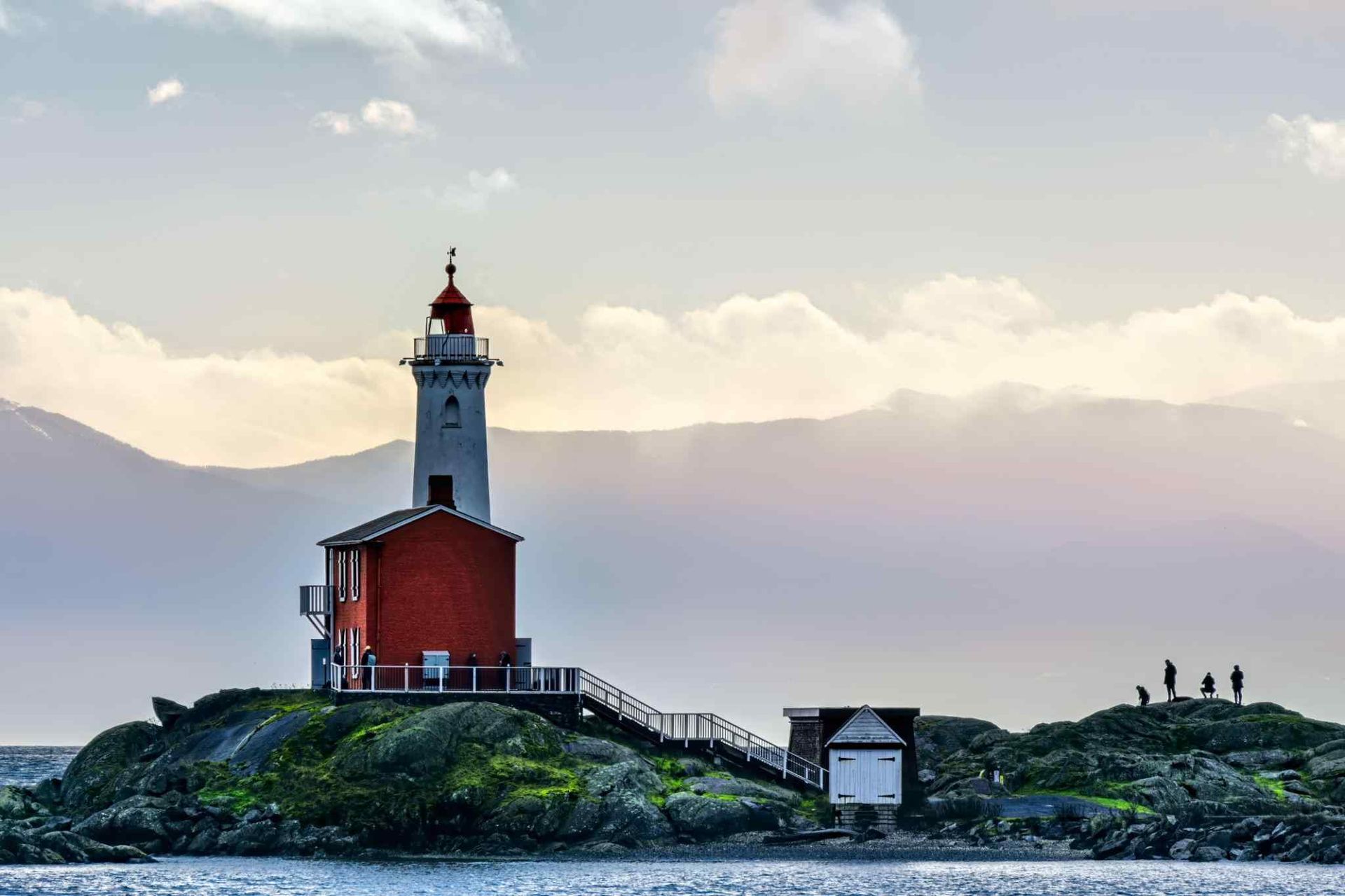 Lighthouse on a rocky shoreline at sunset with a red building and calm water