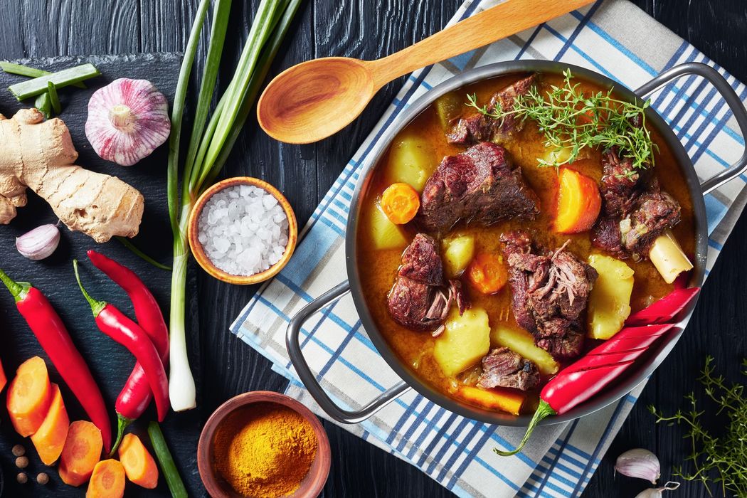Stew with meat, carrots, and potatoes in a pot with ingredients like ginger, garlic, and chili peppers arranged around it.