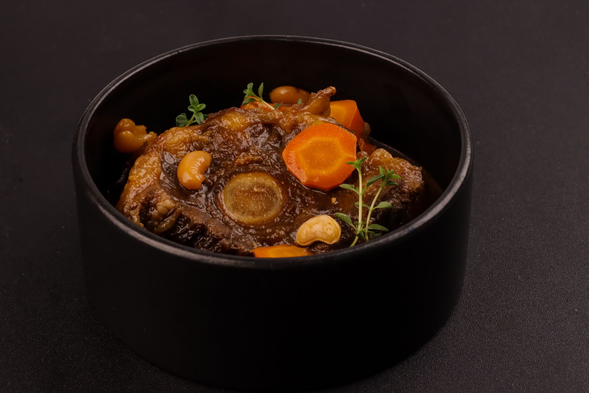 Braised oxtail stew with carrots, herbs, and cashews in a black bowl.