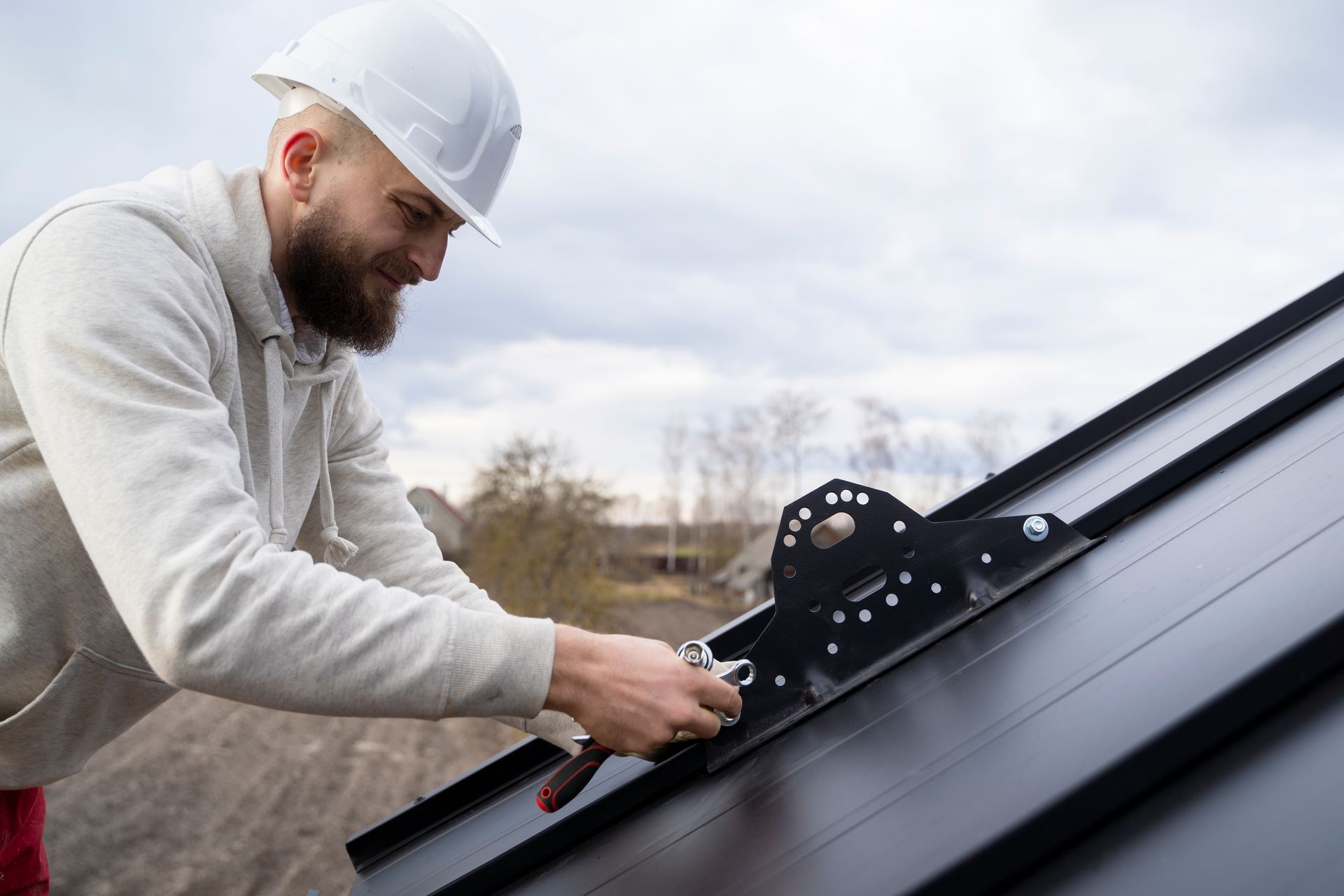 Roofer in white hard hat attaching metal bracket to black roof, working outdoors on cloudy day.