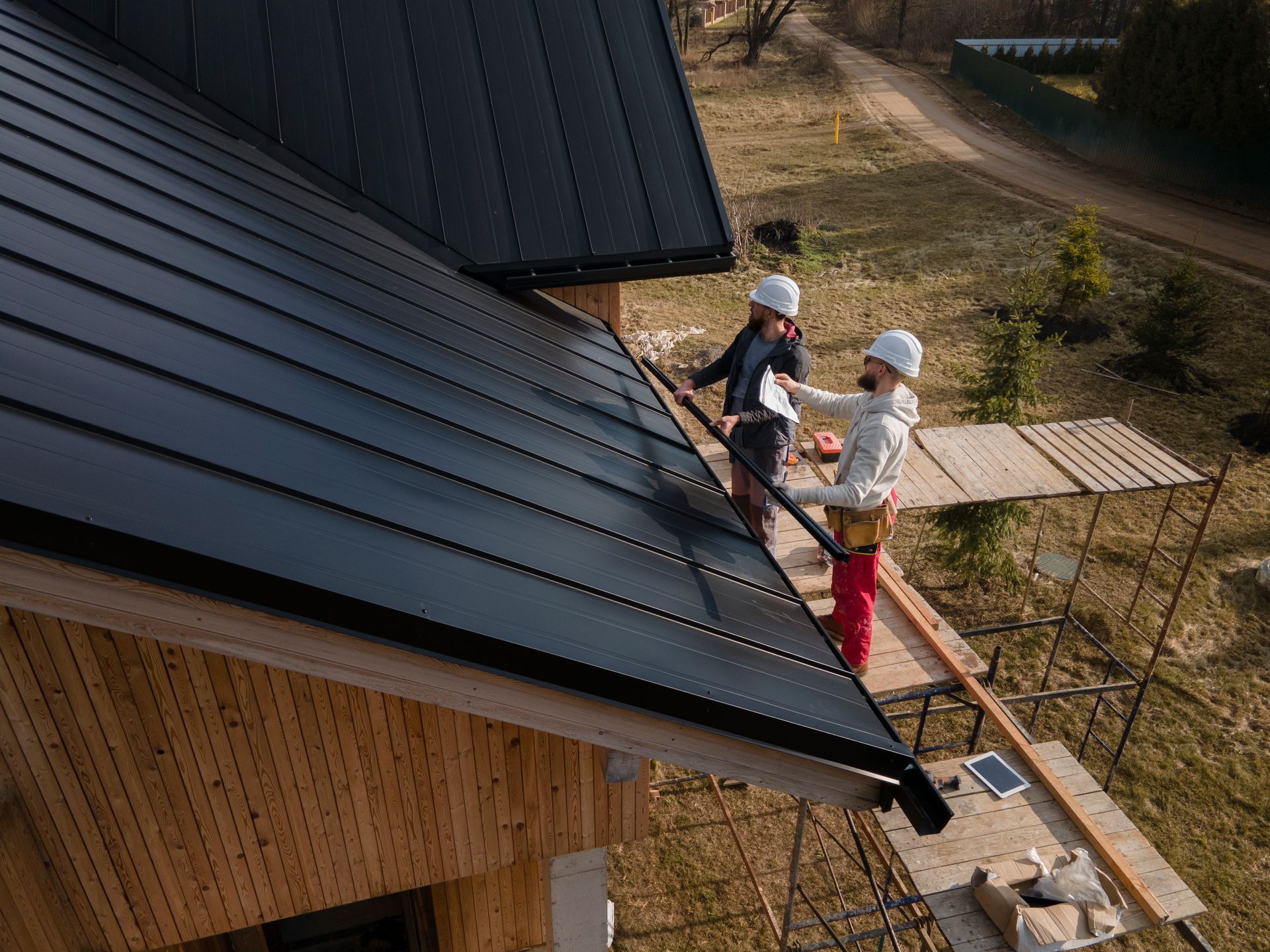 Two roofers in helmets install black metal roofing on a wooden house.