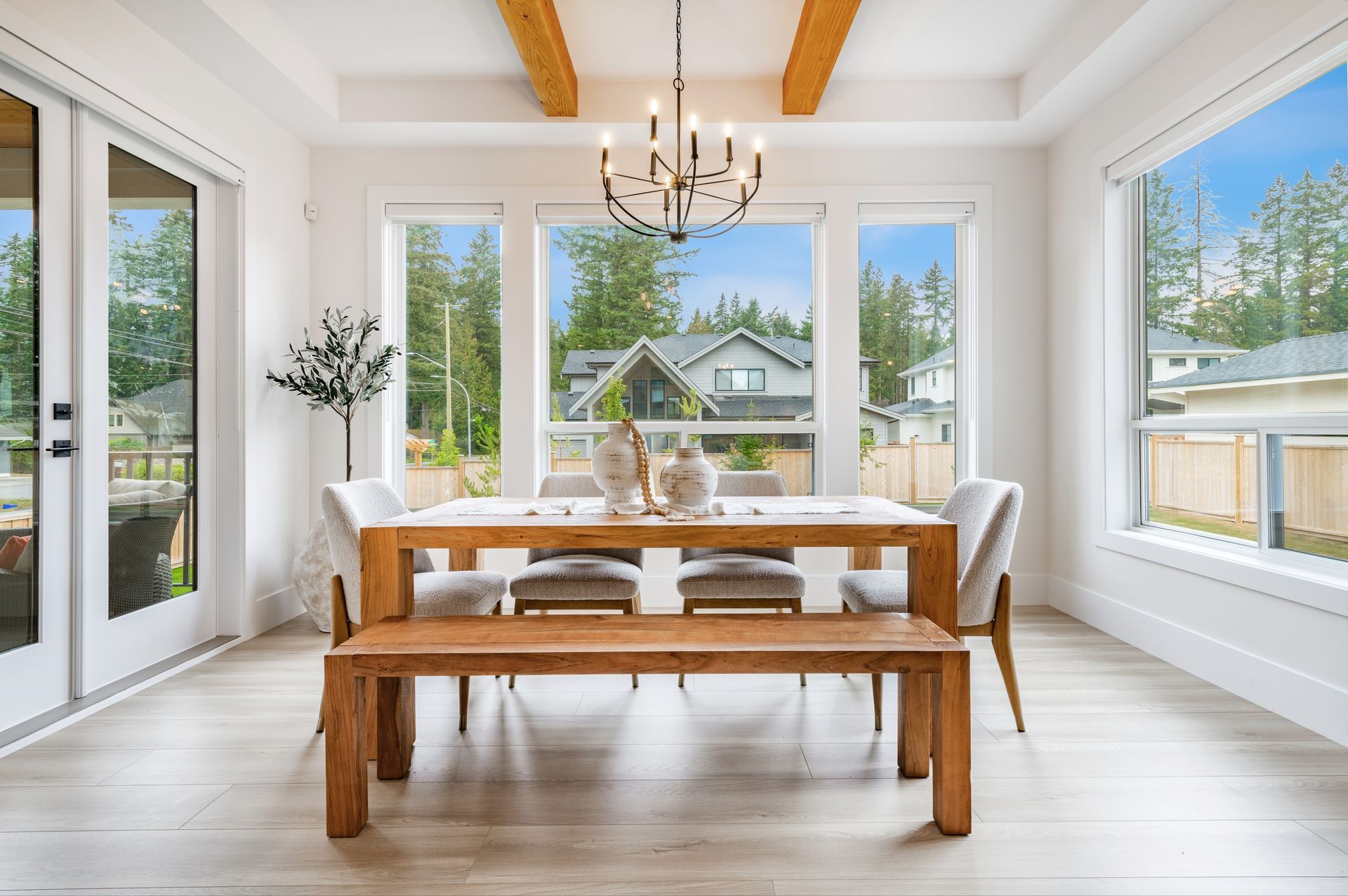 Dining room with wooden table, bench, and chairs, large windows, and chandelier, overlooking a neighborhood.