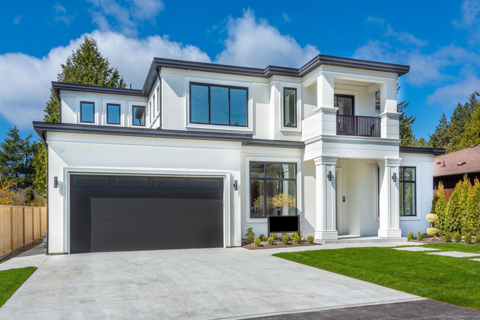 Modern white house with a two-car garage, large windows, balcony, and green lawn on a sunny day.