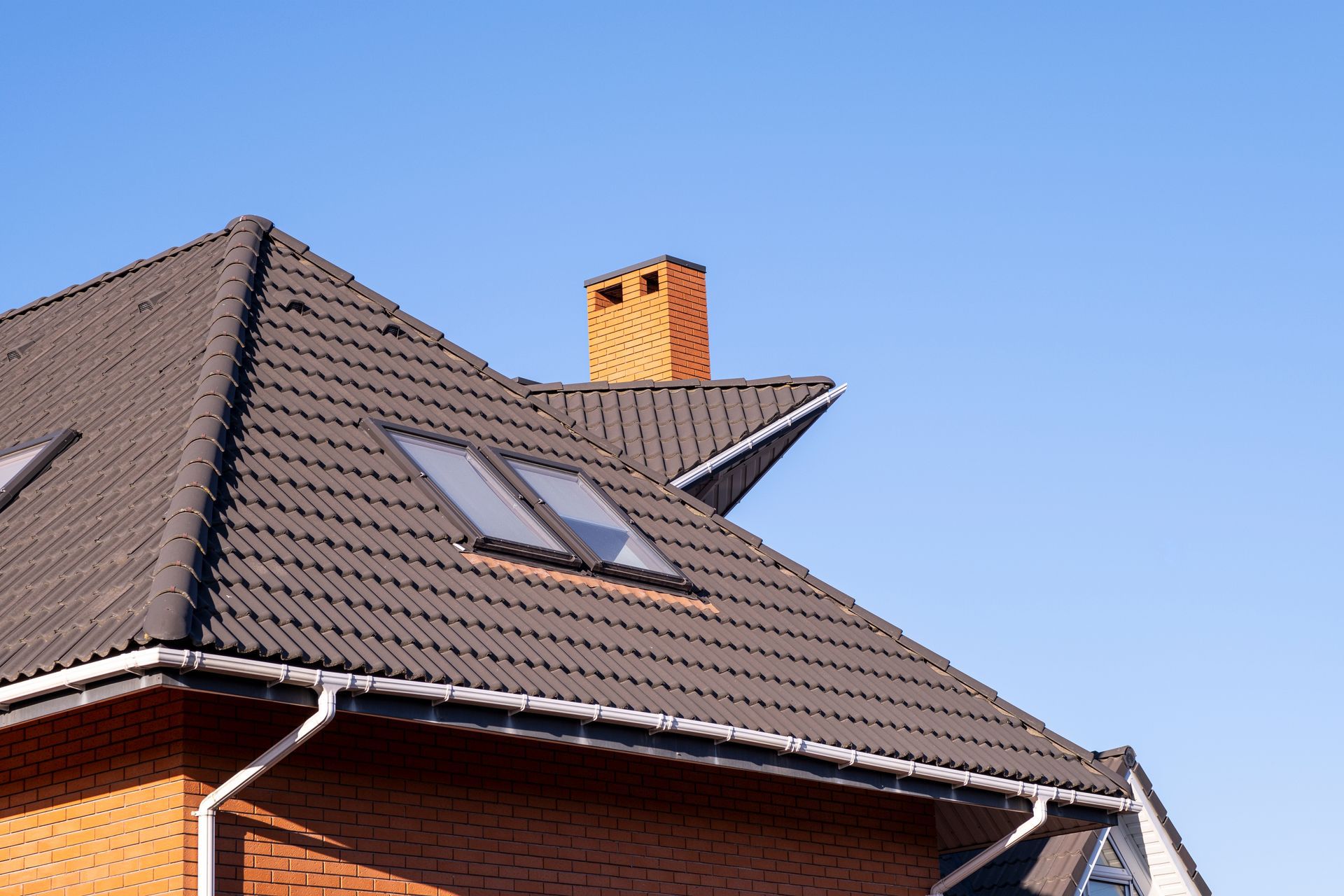 Brown tiled roof with skylights and brick chimney against a blue sky.
