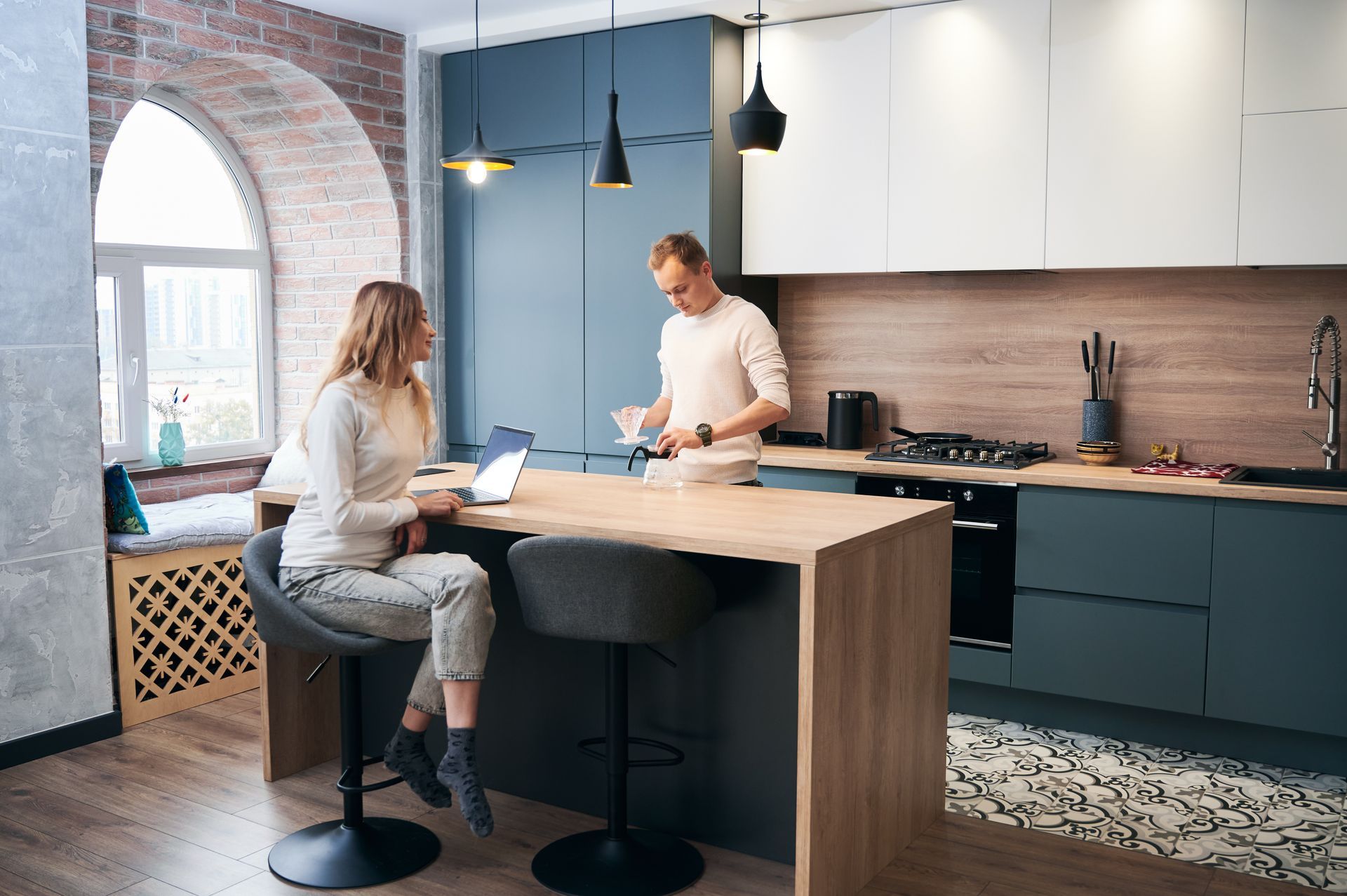 Woman seated at a kitchen island with a laptop; man at the stove. Modern kitchen with blue and wood accents.