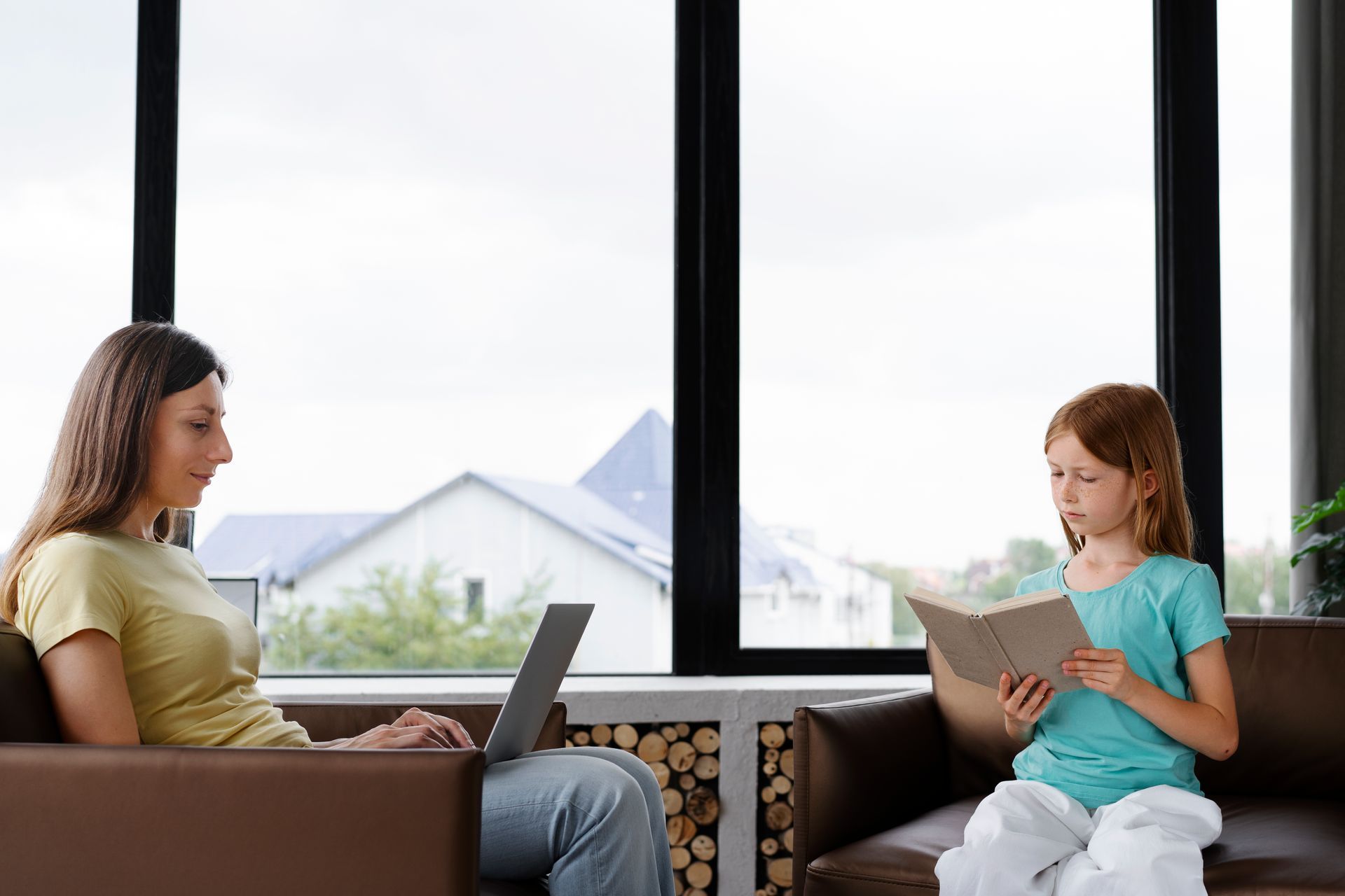 A woman is sitting on a couch using a laptop and a little girl is reading a book.