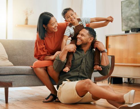 A family of three playfully interacts in a living room. The father sits with the son on his shoulders while the mother looks on smiling.