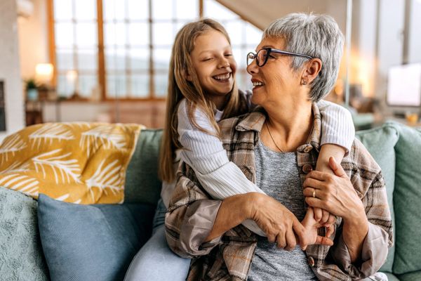 Girl with braces hugs smiling gray-haired woman on a couch indoors. They are looking at each other happily.