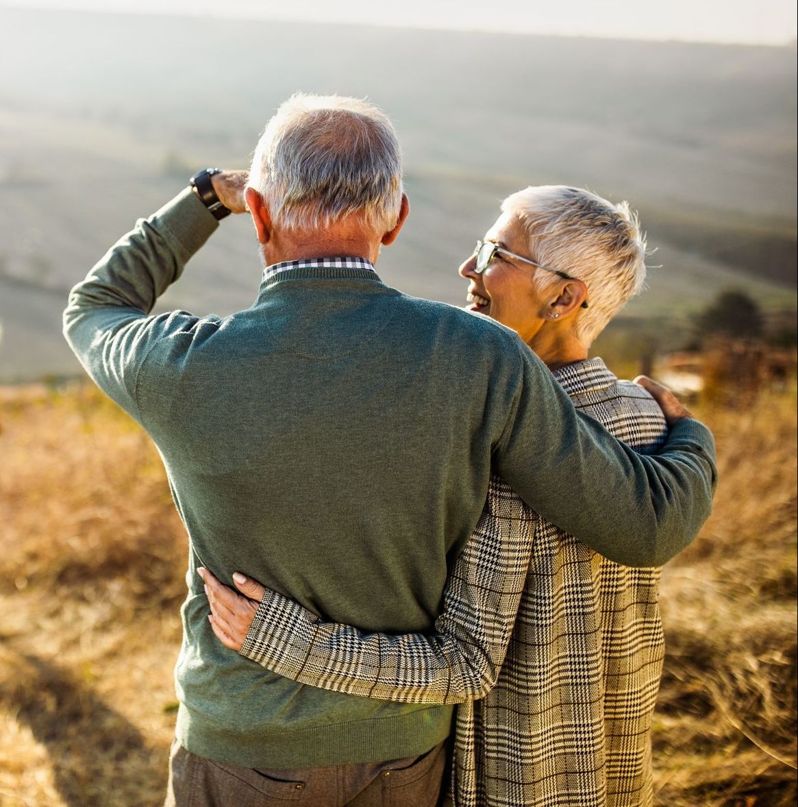 An older couple with gray hair, arms around each other, looking out over a scenic landscape.