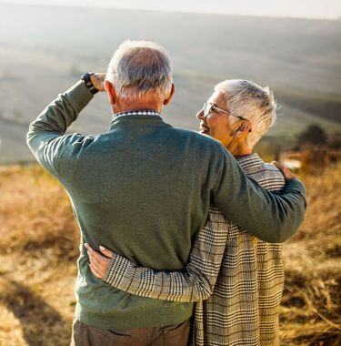 An older couple with gray hair, arms around each other, looking out over a scenic landscape.