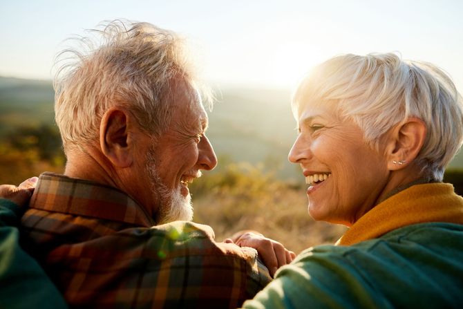 Smiling older couple looking at each other outdoors in a sunny setting. They are embracing, radiating happiness.
