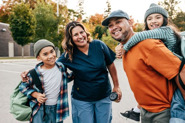 Family of four smiling outdoors: A man carries a girl on his back, while the mom and boy stand beside them.
