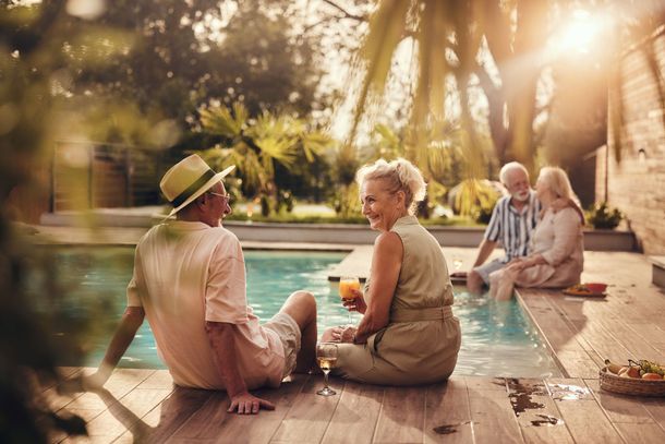 Two couples relaxing by a pool in a sunlit backyard. One couple sits near the pool, smiling, while the other couple sits by the edge.