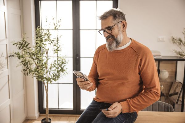 Mature man in orange sweater and glasses smiling at a smartphone; leaning against a table near a window with a plant.