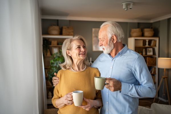 An older couple smiles while holding mugs indoors. The man has his arm around the woman, and they are both looking at each other.
