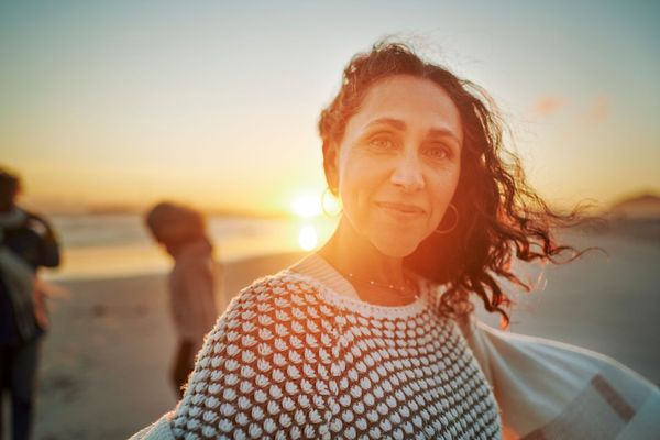 Woman with dark hair and sweater smiles at the camera on a beach at sunset, two people blurred in the background.