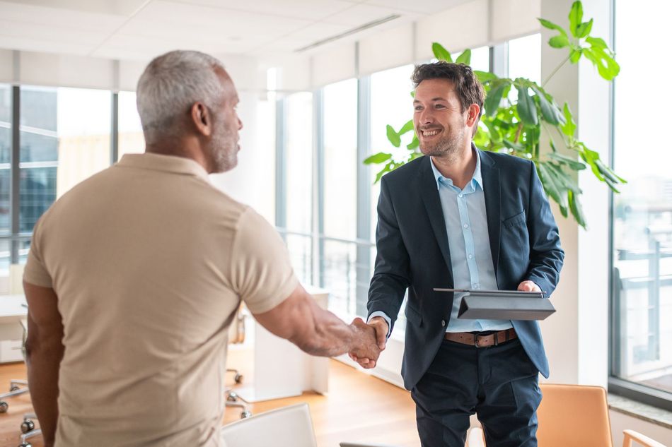 Two men shake hands in an office with a large window and greenery. One man wears a blazer and smiles; the other wears a tan shirt, his back to the camera.