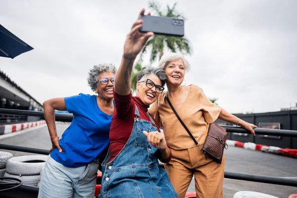 Three smiling older women taking a selfie outdoors, likely at a go-kart track. One holds the phone, the others pose.