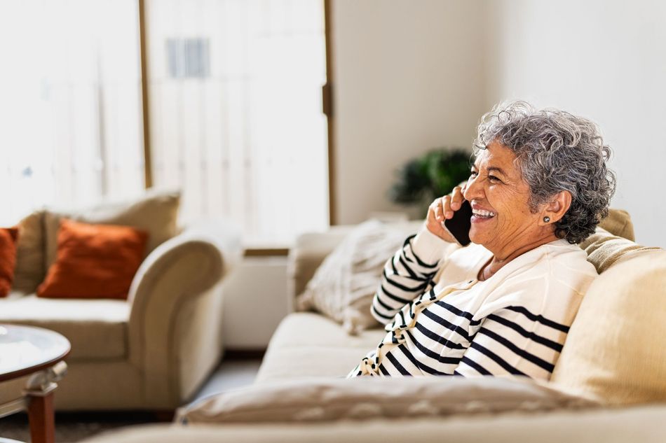 An elderly woman with gray curly hair smiles while talking on a cell phone. She sits on a beige couch in a living room.
