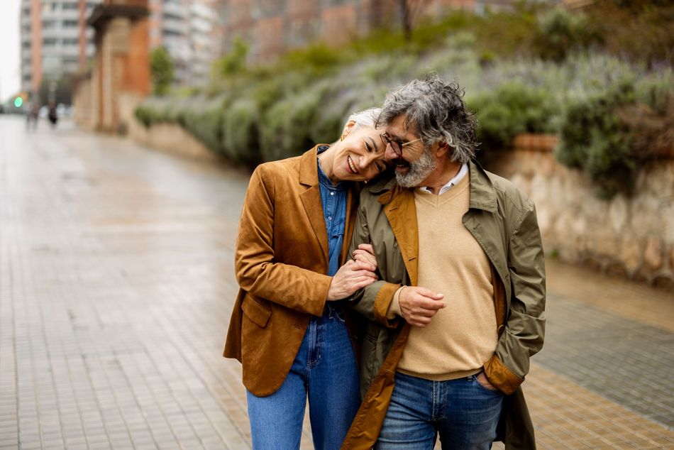 An older couple walking arm-in-arm on a city street; woman resting head on man's shoulder, both smiling.
