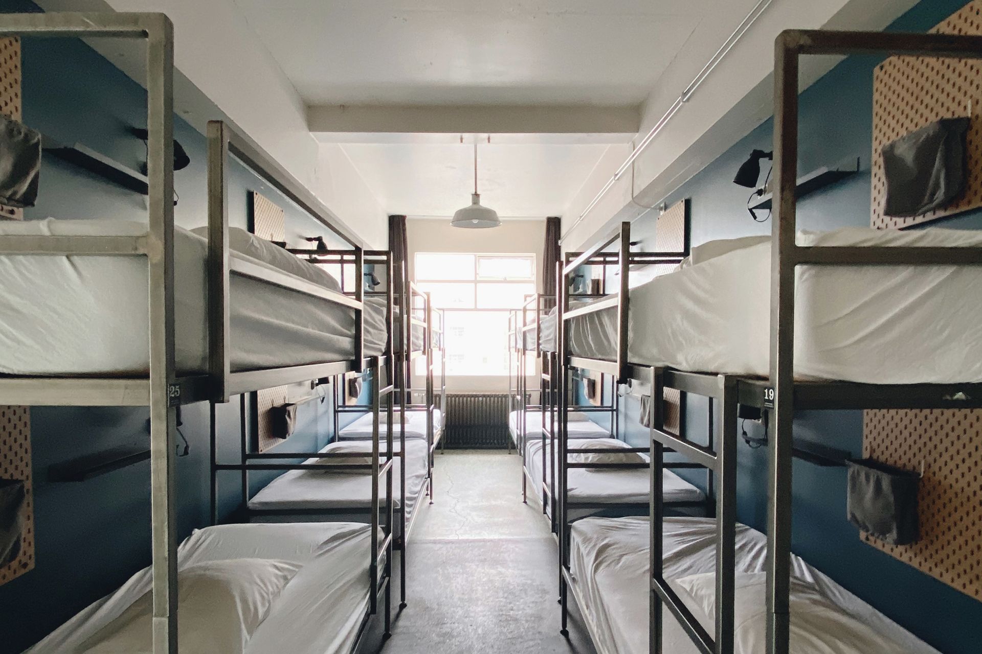Bunk beds line a narrow hostel room. White sheets, dark metal frames, and blue walls. Window at end.