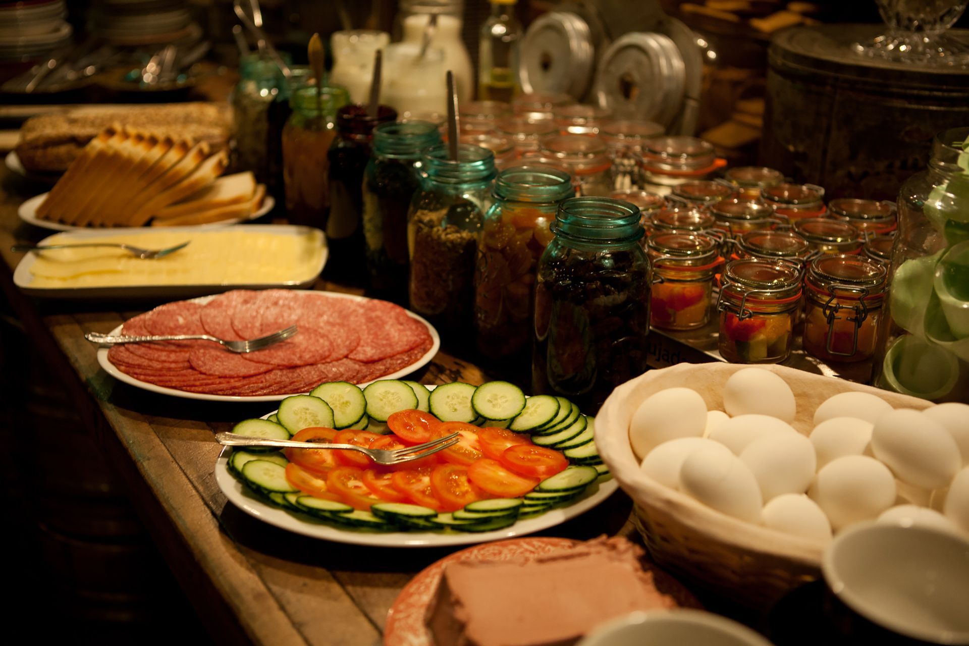 Breakfast buffet: cold cuts, sliced vegetables, eggs, cheeses, and various jams in jars.