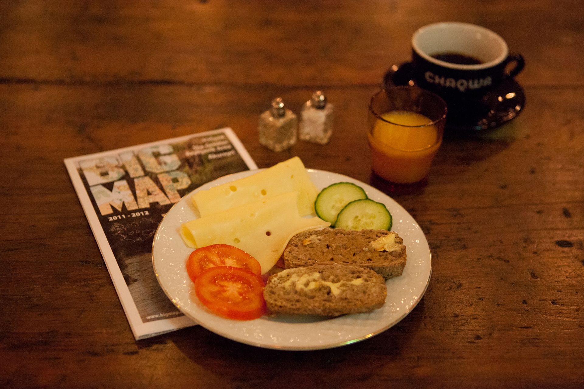 Breakfast plate with cheese, bread, tomato, and cucumber; coffee, orange juice, and a magazine on a wooden table.