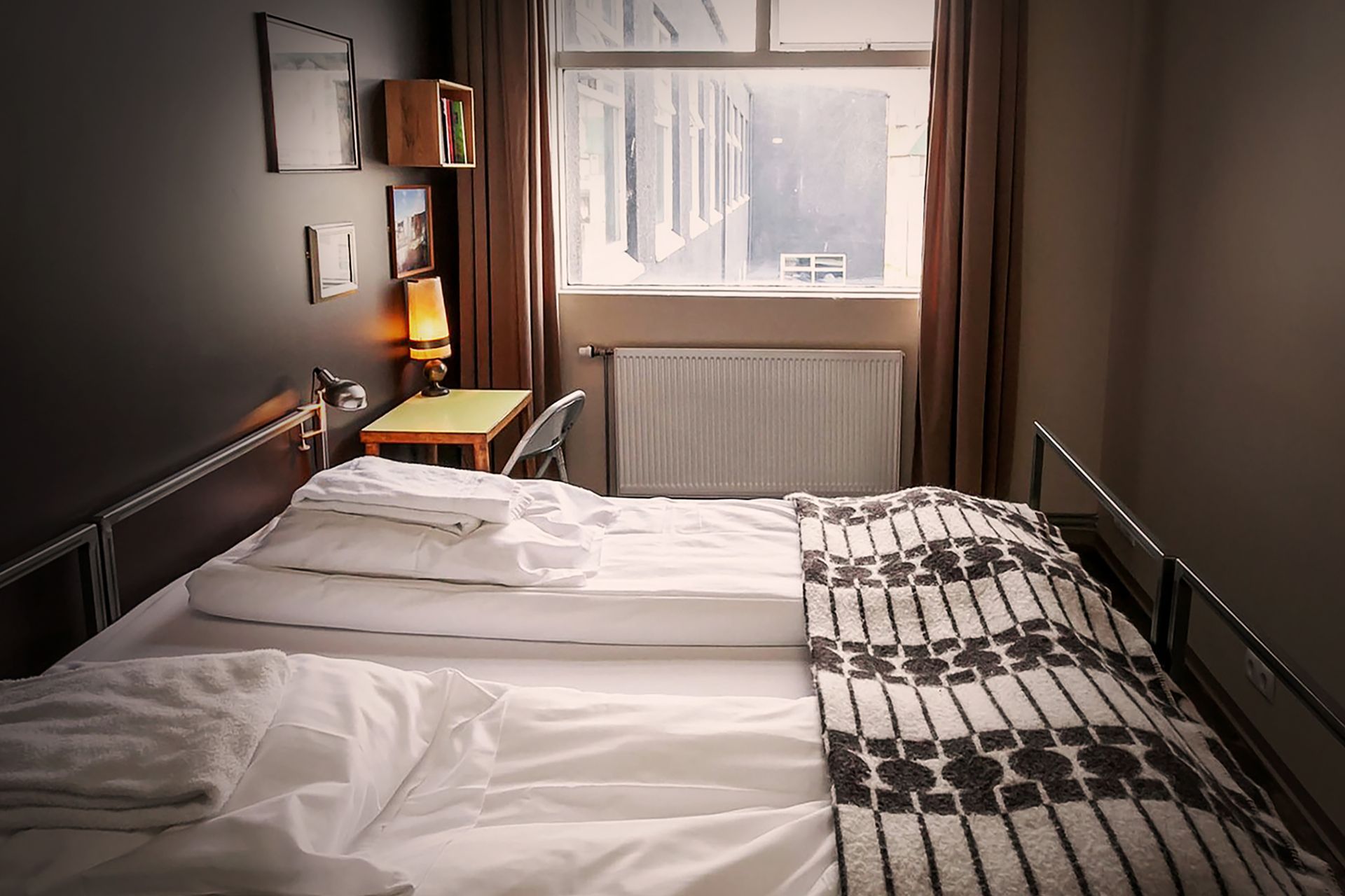 Bedroom with unmade bed, small desk by window, dark walls, warm lighting.