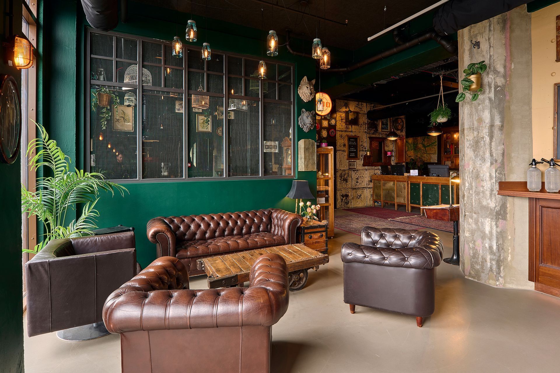 Leather sofas in a dimly lit, vintage-style lounge with dark green walls, a large window, and decorative lighting.