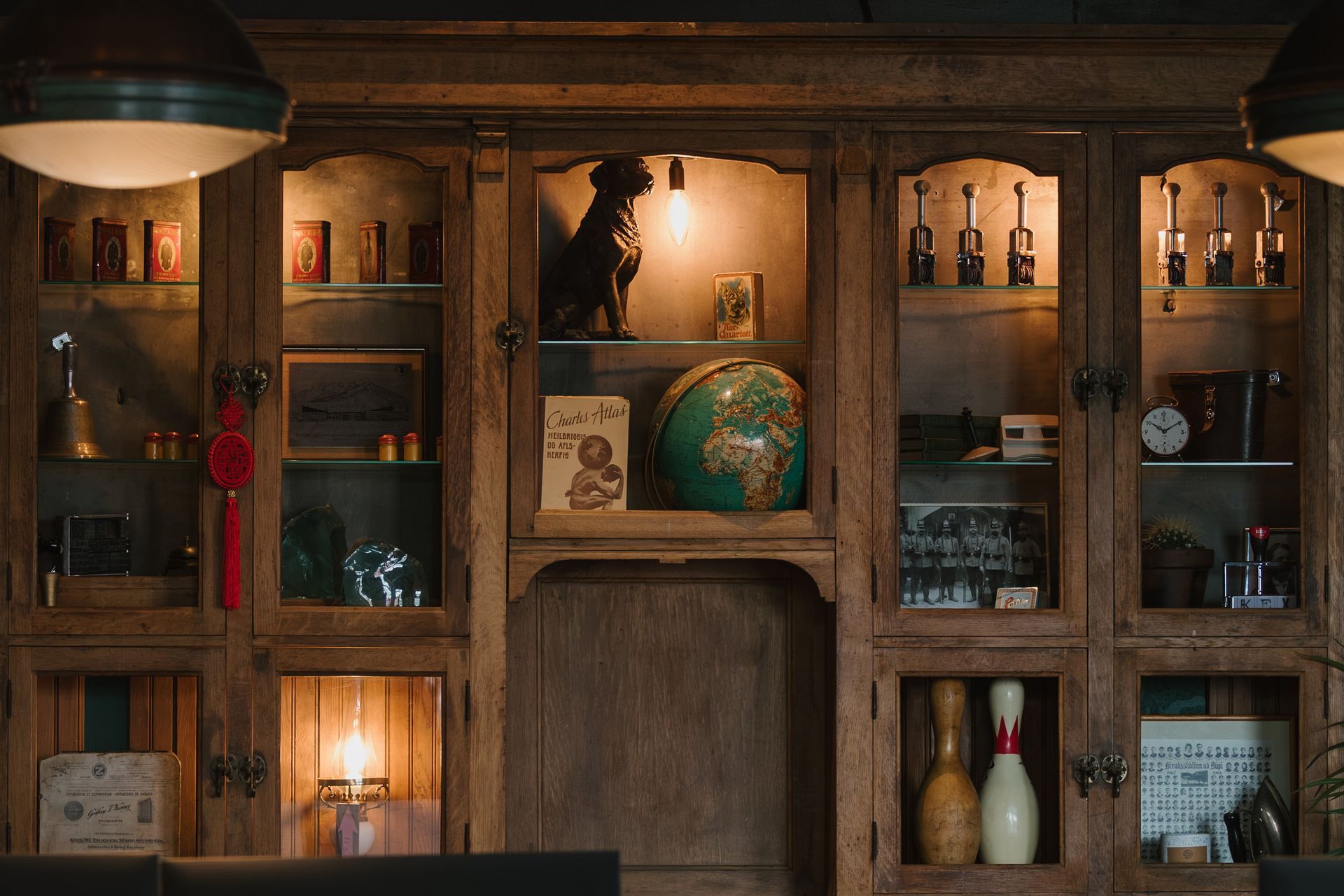 Wooden cabinet filled with various items, including a dog statue, globe, and bowling pins, illuminated by interior lights.