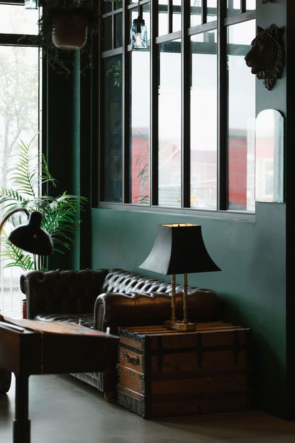 Dark green-walled lounge with leather sofa, trunk side table, lamp, and window view of a red brick building.